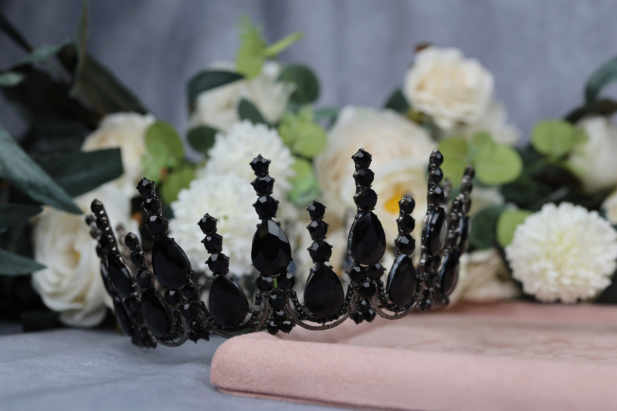 A black ornate crown or tiara with black gemstones is placed on a pink surface, surrounded by white flowers.