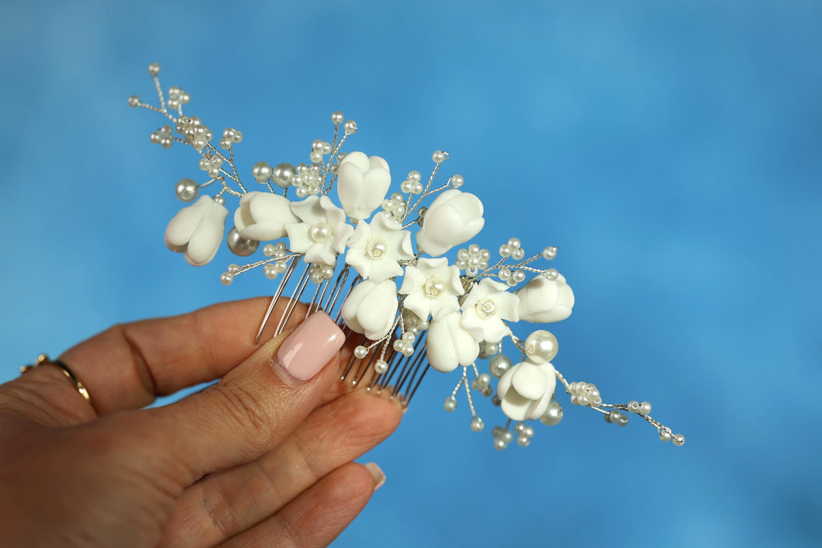 A hand holding a decorative hair clip with white flowers and pearls against a blue background.