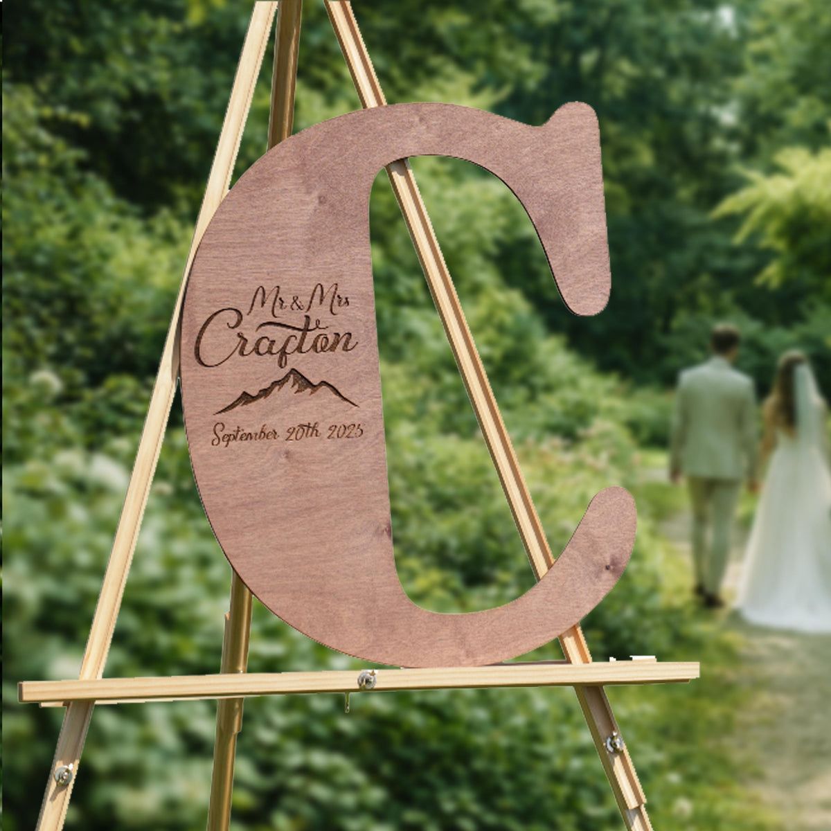 A wooden letter 'C' with the text "Mr. & Mrs. Crafton September 20th 2023" engraved on it, standing on a wooden easel in a garden setting.