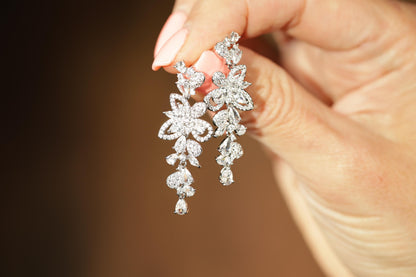 A hand holding a pair of elegant diamond earrings against a blurred background.