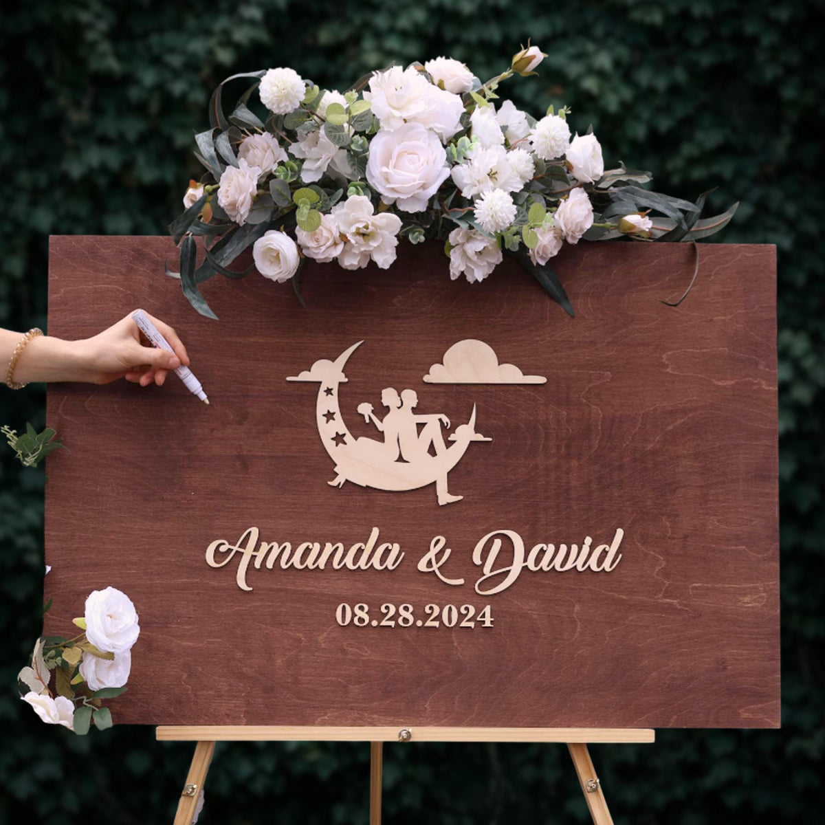 A wooden sign with a floral arrangement and a silhouette of a couple, along with the text "Amanda & David" and the date "08.28.2024".