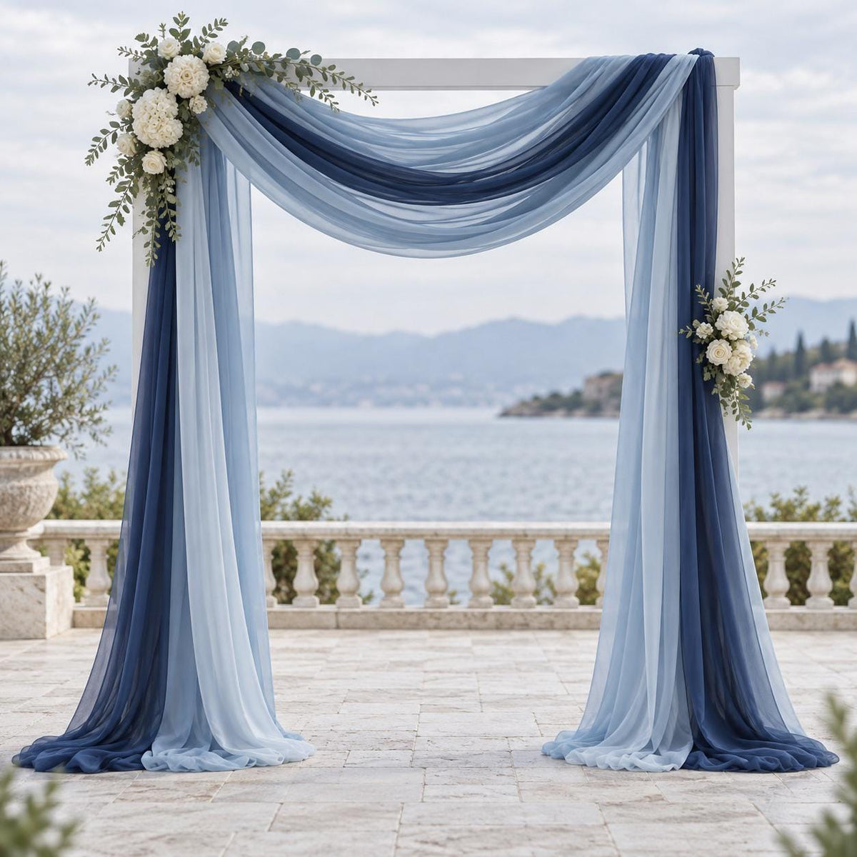 a beautiful wedding arch adorned with blue and white fabric, flowers, and greenery, set against a scenic backdrop of a body of water and mountains.