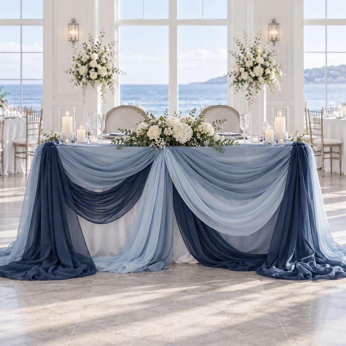 a beautifully decorated wedding reception table with a blue and white color scheme. The table is adorned with white flowers and draped in blue fabric, creating a romantic and elegant atmosphere. The table is set with candles, plates, and silverware, suggesting a formal dining experience. The table is positioned in front of a large window, offering a scenic view of the ocean.