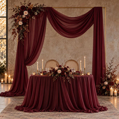 a beautifully decorated wedding reception table with a red tablecloth, floral centerpieces, and candles. The table is set for a formal event, with a large floral arch and a draped curtain framing the table. The overall setting has a romantic and elegant atmosphere.