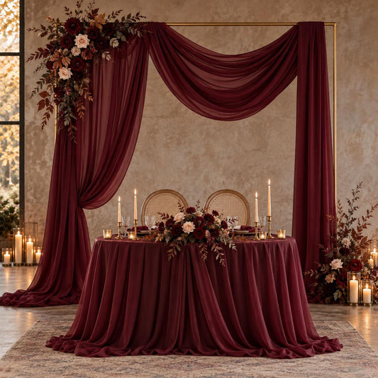 a beautifully decorated wedding reception table with a red tablecloth, floral centerpieces, and candles. The table is set for a formal event, with a large floral arch and a draped curtain framing the table. The overall setting has a romantic and elegant atmosphere.