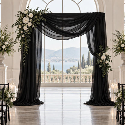 a beautifully decorated wedding arch with black fabric and white flowers, set against a backdrop of a large window overlooking a scenic view of mountains and a body of water.