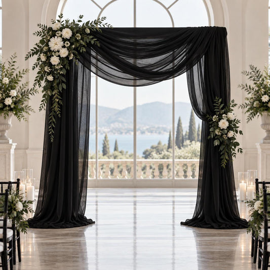 a beautifully decorated wedding arch with black fabric and white flowers, set against a backdrop of a large window overlooking a scenic view of mountains and a body of water.