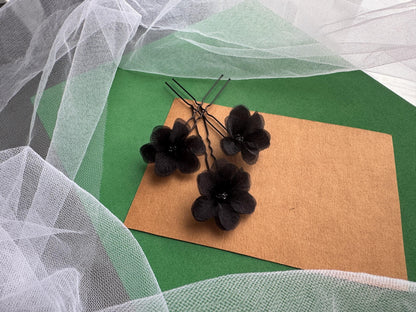 a close-up view of black flower-shaped hair clips and a brown paper card on a green surface, with a white net or veil in the background.