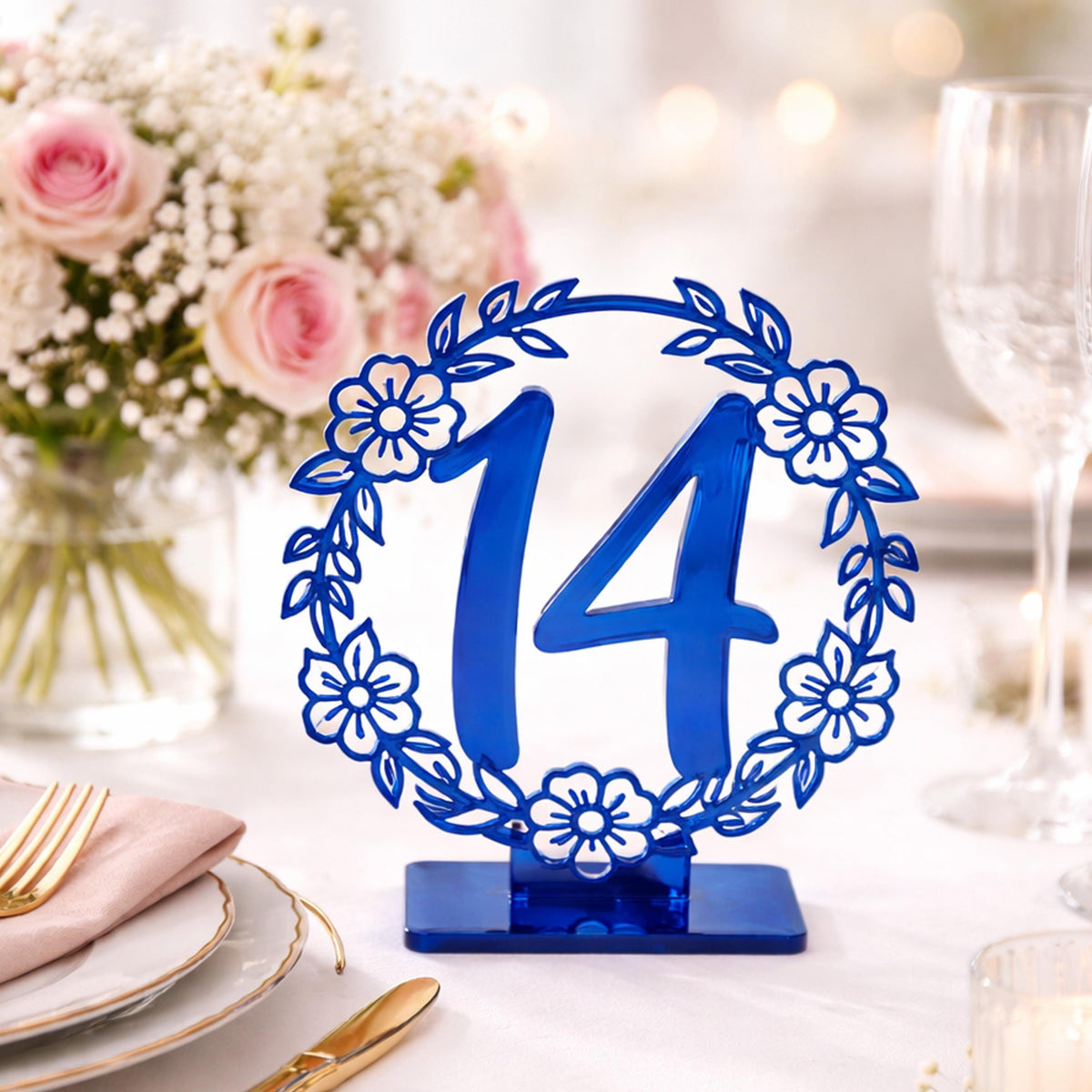 A blue table number holder with the number 14 in the center, surrounded by a floral design, sits on a table with a vase of pink roses and white flowers in the background.