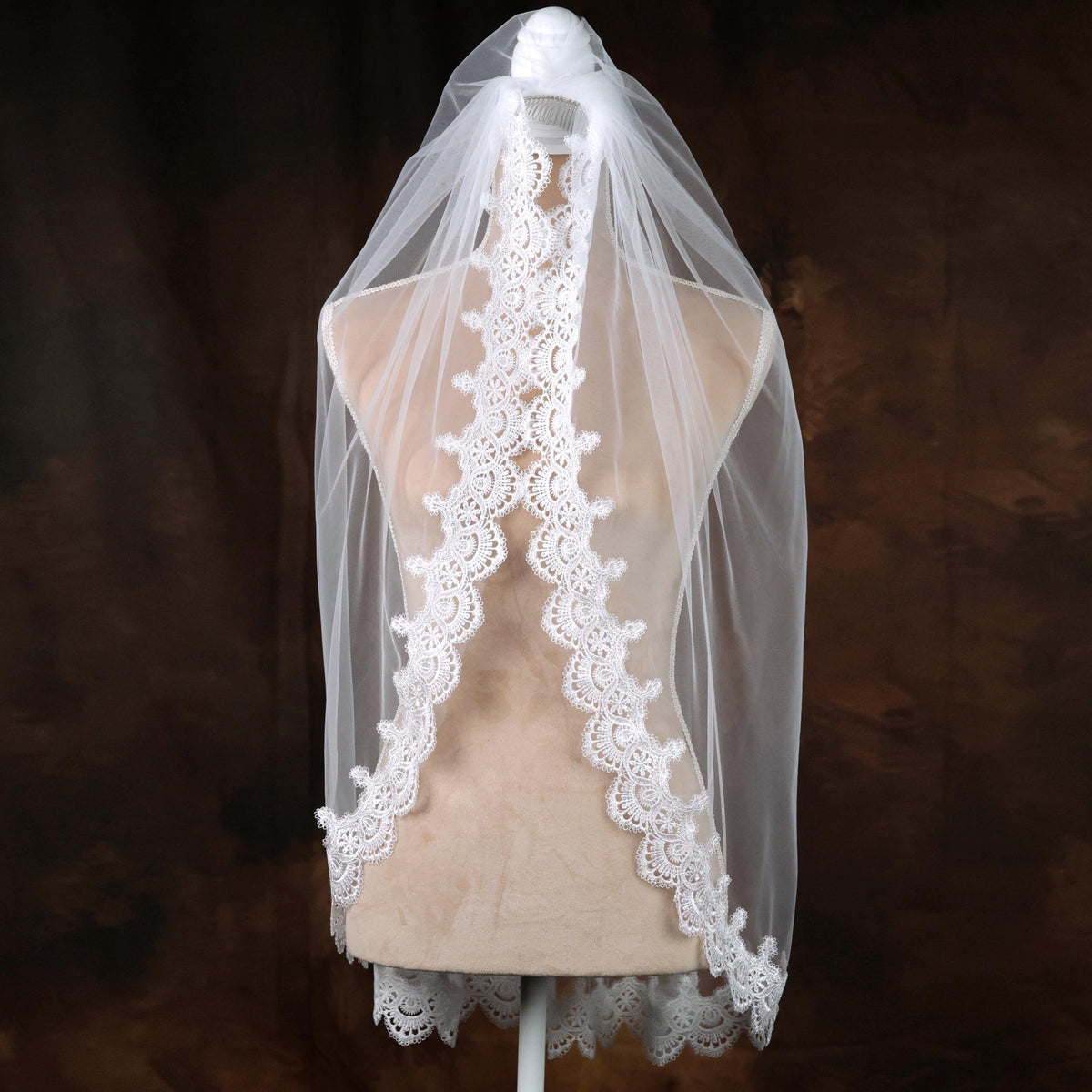 A mannequin wearing a white lace veil with a scalloped edge, displayed against a dark background.