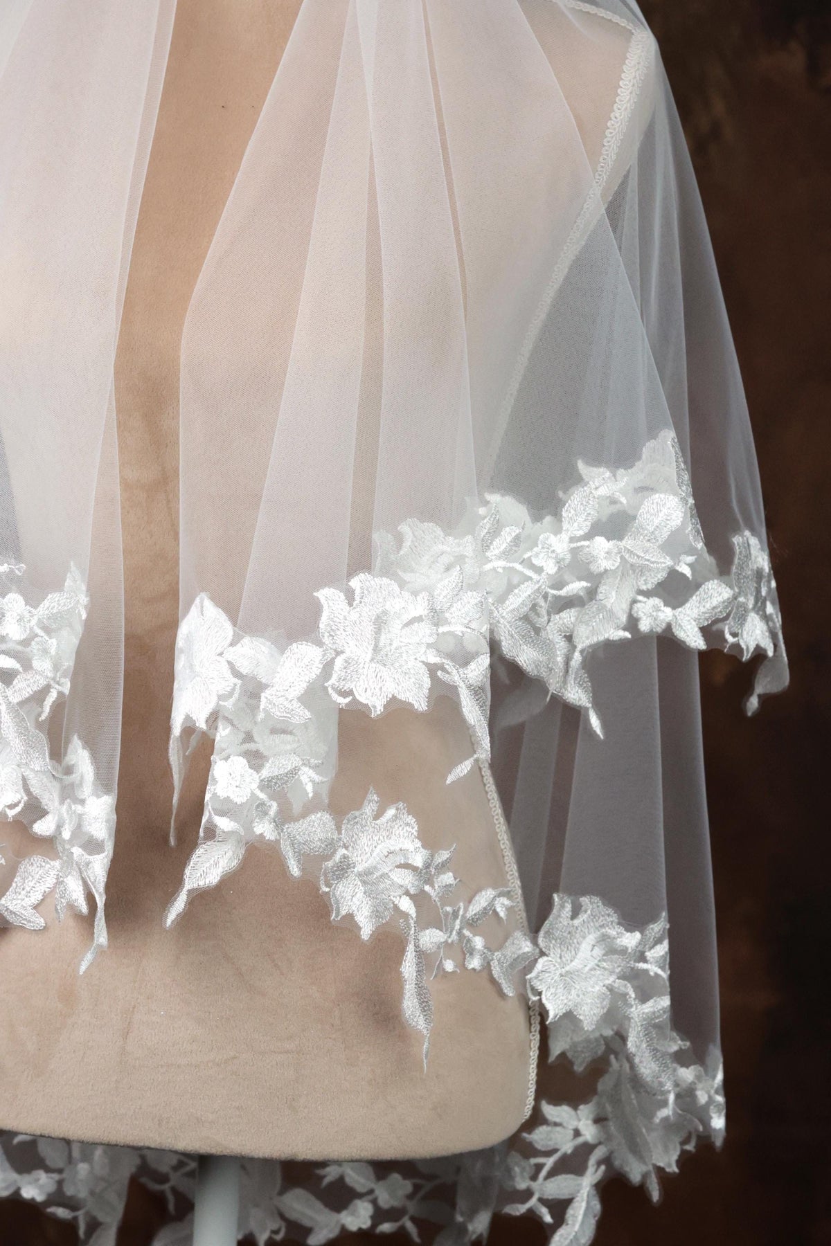 A close-up view of a delicate white lace veil with intricate floral embroidery, displayed on a mannequin or dummy.