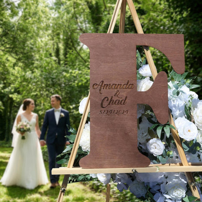A bride and groom walking hand in hand in a lush green garden, with a large wooden letter 'F' featuring the names 'Amanda & Chad' and the date '9/23/2021' in the foreground.