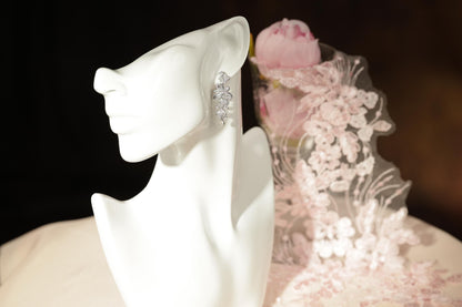 A white mannequin head wearing a pair of earrings, with a pink flower in the background.