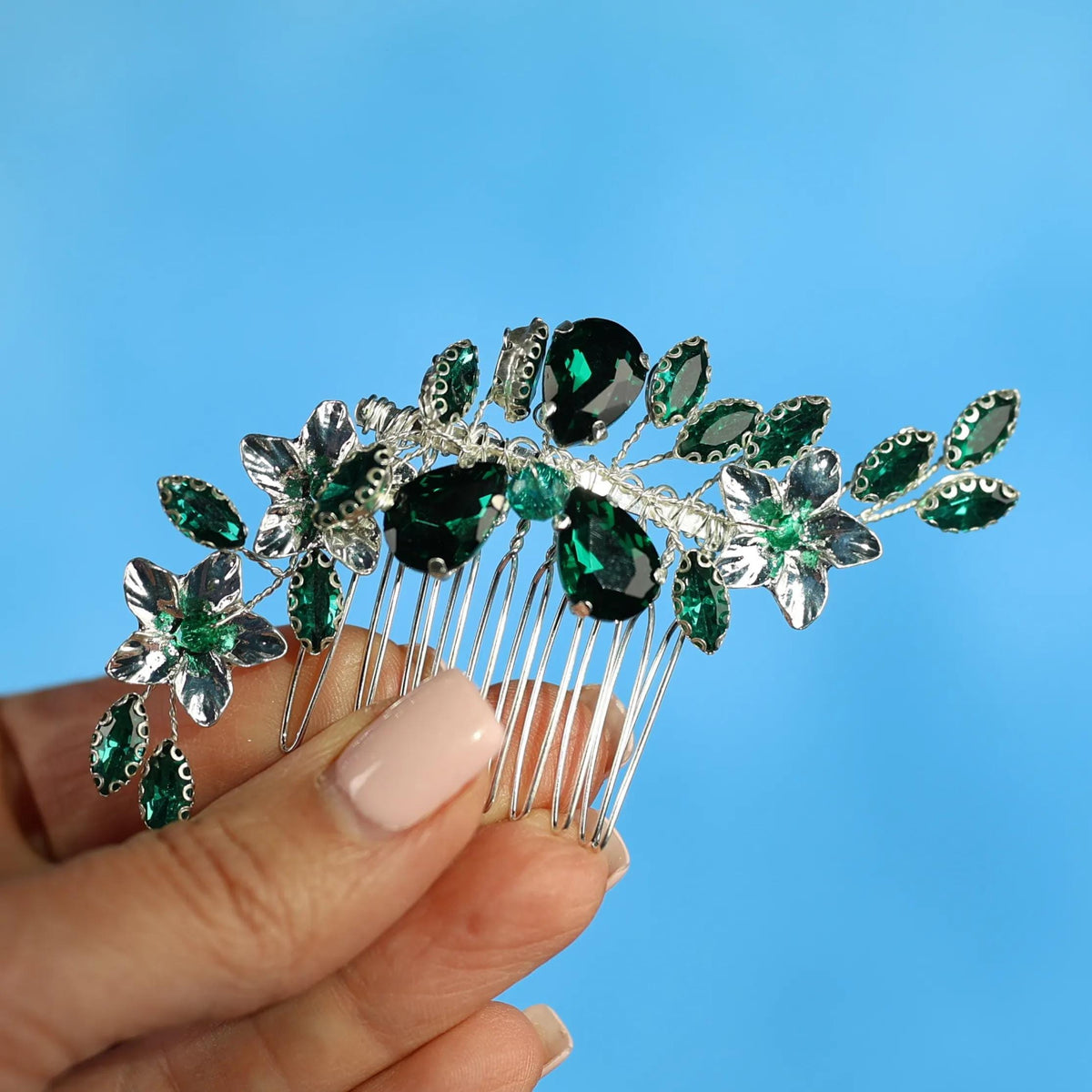 A hand holding a decorative hair accessory with green and silver gemstones against a blue background.