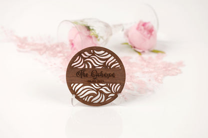 A wooden coaster with a zebra print design and the name "The Johnson" engraved on it, placed on a white surface with a glass of wine and a pink rose in the background.