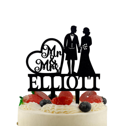 A black silhouette of a bride and groom on a wedding cake topper, with the name "Ellott" written below them.