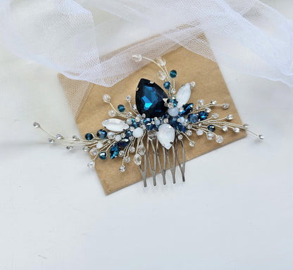 A decorative hair accessory with a large blue gemstone and smaller blue and white gemstones, as well as silver and white beads, is displayed on a white surface.