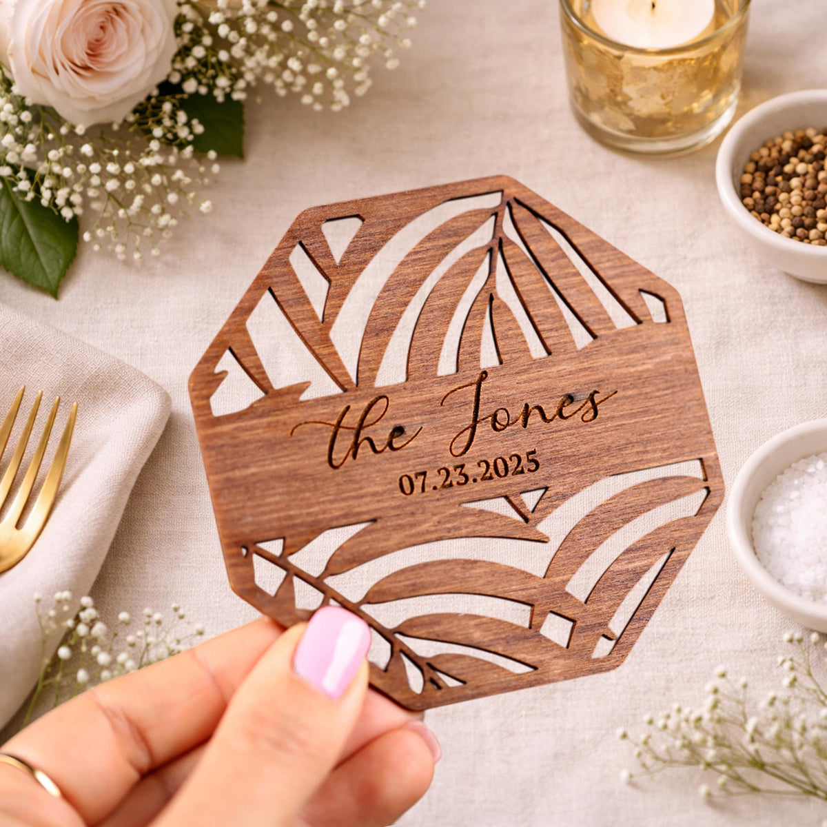 A hand holding a wooden coaster with a palm tree design, surrounded by other decorative items such as a rose, a candle, and a bowl of spices.