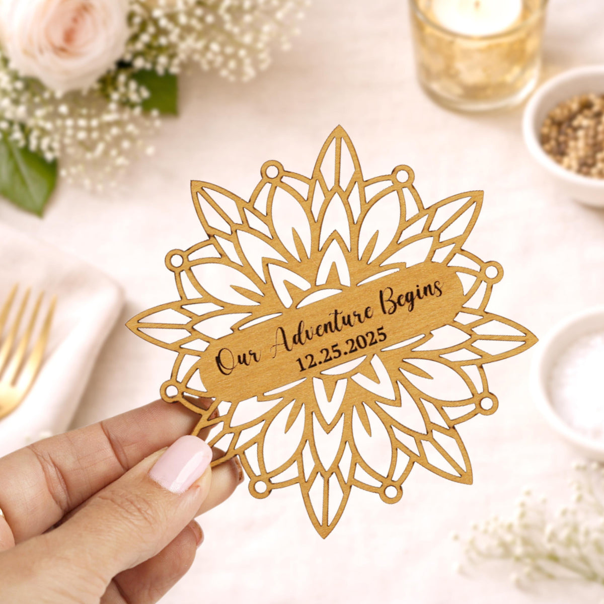 A hand holding a wooden ornament with the text "Our Adventure Begins 12.25.25" on it, surrounded by flowers and other decorative elements.
