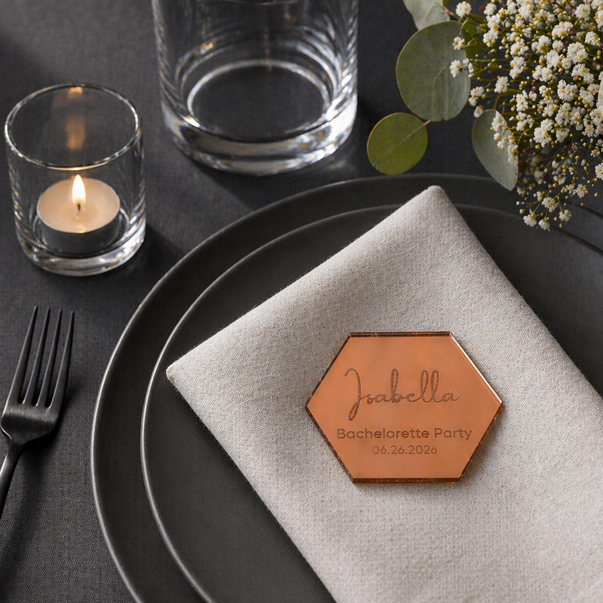a table setting with a plate, a candle, a glass, and a floral arrangement. There is a hexagonal label on the napkin that says "Joabella Bachelorette Party 06.26.2026".