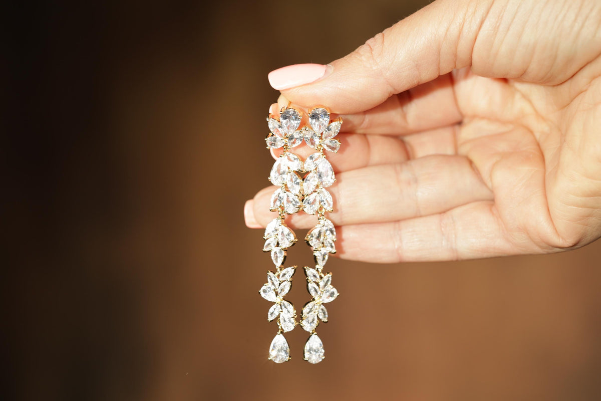 A hand holding a pair of elegant diamond earrings against a blurred background.