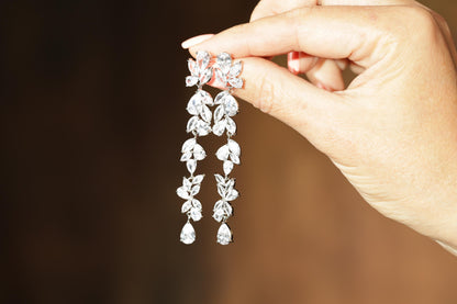 A hand holding a pair of diamond earrings with multiple dangling crystal elements.