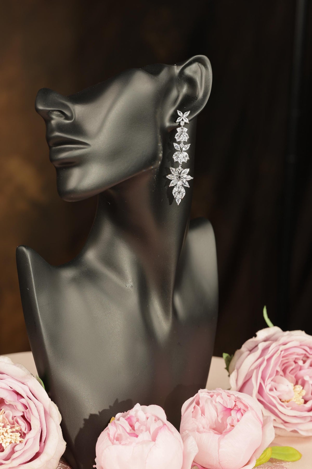 A black mannequin head with a necklace featuring white and pink flowers, set against a dark background with pink peonies.