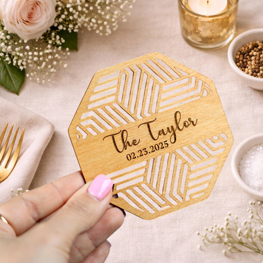 A hand holding a wooden coaster with the name "The Taylor" and the date "02.25.2025" printed on it, surrounded by other decorative items such as a rose, a candle, and some spices.