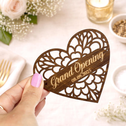 A wooden heart-shaped card with the text "Grand Opening 08.09.2025" is being held in a hand, with a floral arrangement and other decorative items visible in the background.