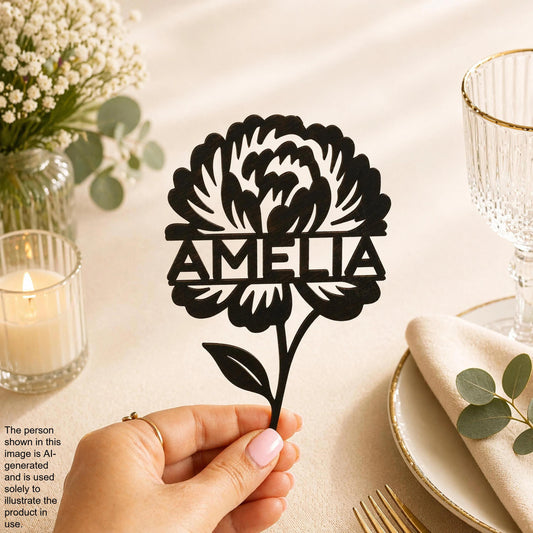 A hand holding a black paper cutout of a flower with the name "Amalia" written on it, placed on a table with a candle, glass, and other decorative items.