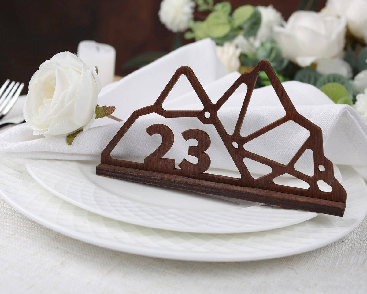 A wooden place card holder with the number 23 on it, placed on a white plate, with a white rose and a fork nearby.