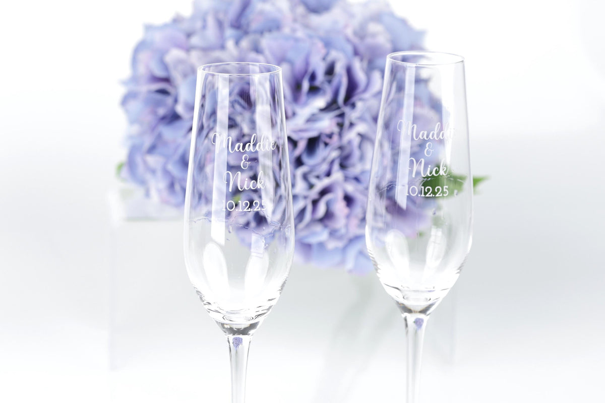 two champagne glasses with personalized messages, placed in front of a bouquet of purple flowers.