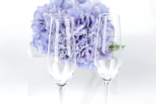 two champagne glasses with personalized messages, placed in front of a bouquet of purple flowers.