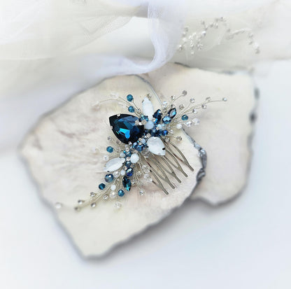 A decorative hair clip with a blue gemstone and white and blue crystal embellishments rests on a white surface.
