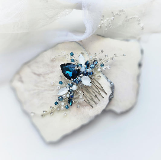 A decorative hair clip with a blue gemstone and white and blue crystal embellishments rests on a white surface.
