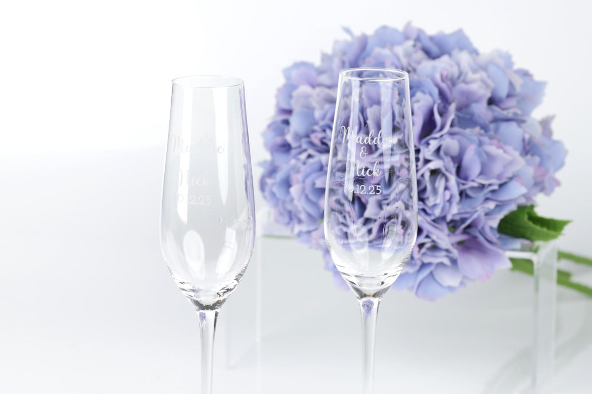 two empty champagne flutes with personalized messages, placed in front of a bouquet of purple flowers.