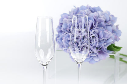two empty champagne flutes with personalized messages, placed in front of a bouquet of purple flowers.