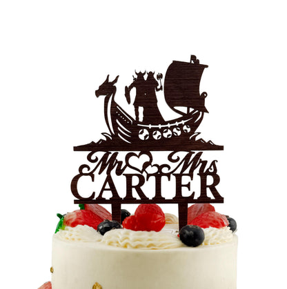 A cake with a wooden topper featuring a silhouette of a couple and a ship, along with the name "Mr. Carter" written on it.