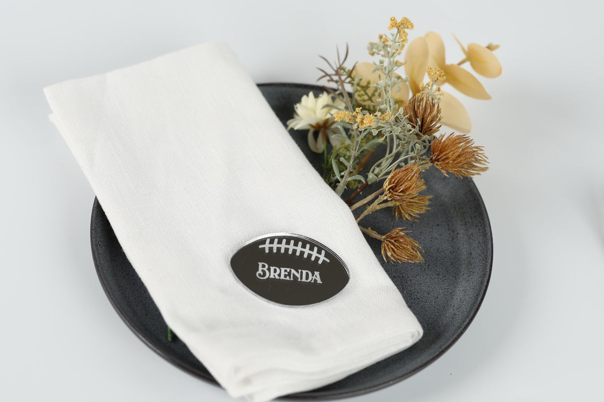 A white napkin with a black football-shaped name tag on it, placed on a black plate with a small arrangement of yellow flowers.