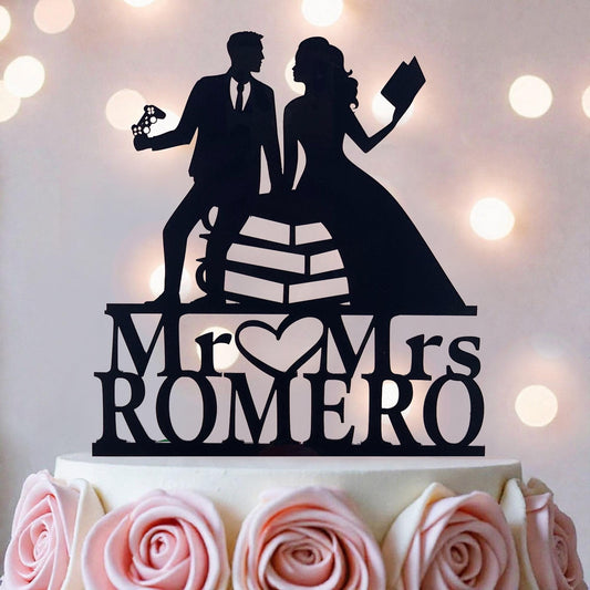 A silhouette of a man and a woman holding a book and a remote control, standing on a cake topper that says "Mr. & Mrs. ROMERO".