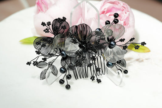 A decorative hair accessory with black and white floral elements, including leaves and pearls, is displayed on a white surface.