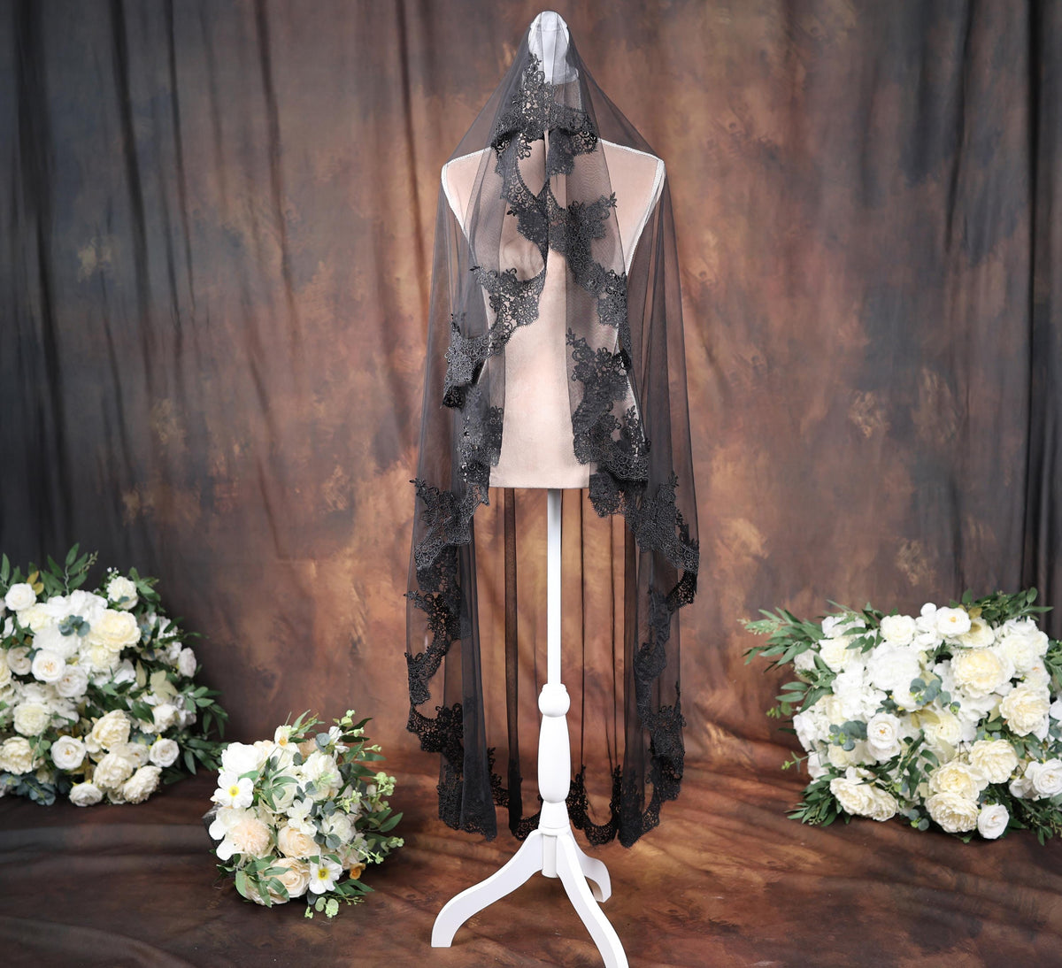 a mannequin wearing a black lace veil, surrounded by white flowers and a brown curtain backdrop.