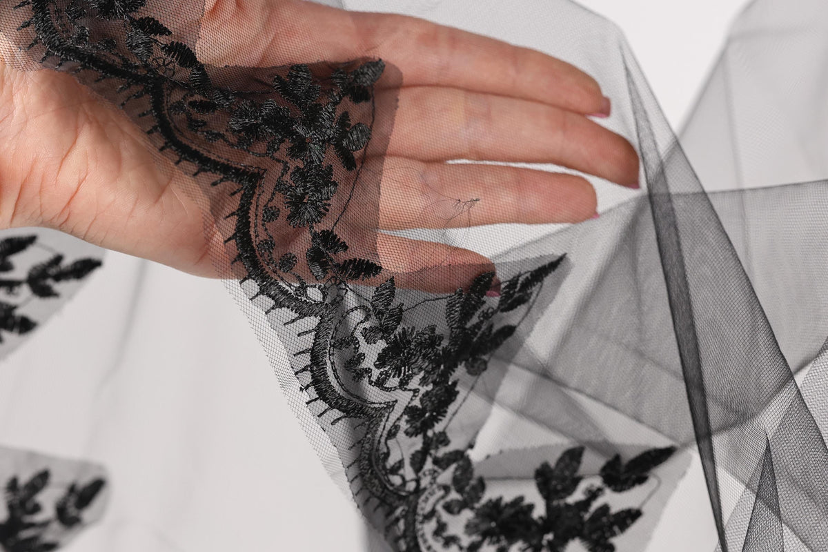 A hand is holding a piece of black lace with intricate floral embroidery.
