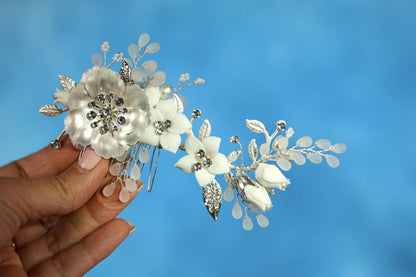 A hand holding a decorative hair accessory with white flowers and silver embellishments against a blue background.