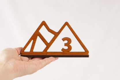 A hand is holding a wooden object that resembles a mountain or mountain-like shape, with the number "3" prominently displayed in the center.