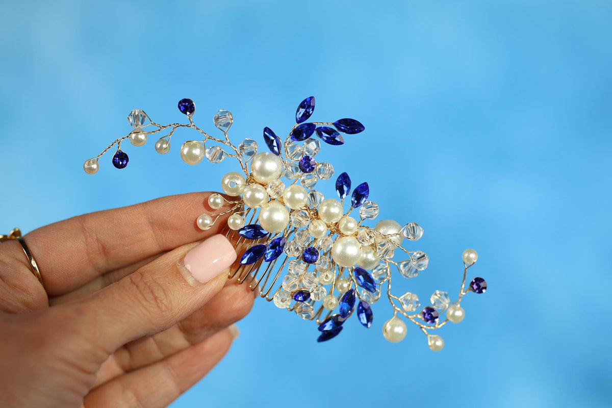 A hand holding a decorative hair accessory with blue and white elements, including pearls and crystals.