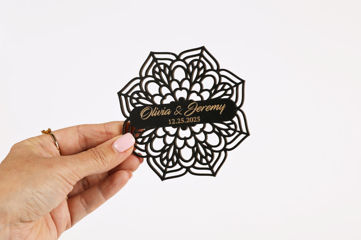 A hand holding a black, ornate paper with the names 'Olivia & Jeremy' and the date '12.25.2025' printed on it.