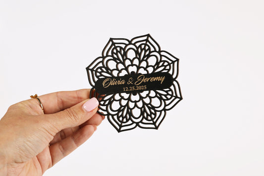 A hand holding a black, ornate paper with the names 'Olivia & Jeremy' and the date '12.25.2025' printed on it.