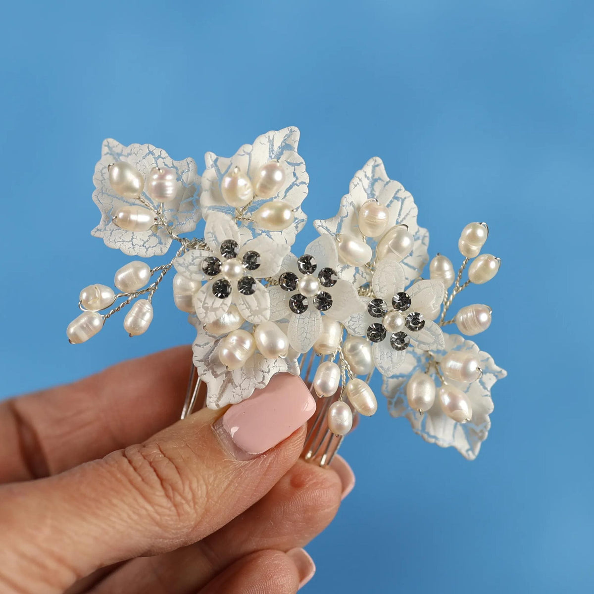 A hand holding a decorative hair accessory with white leaves and pearls against a blue background.