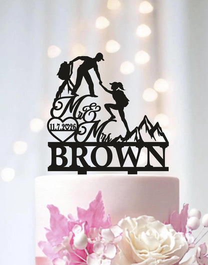 A black silhouette of a couple climbing a mountain on a cake topper, with the text "Mr & Mrs BROWN" below it.
